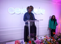 A woman in front of a "celebrate" neon sign and behind a podium addressing attendees