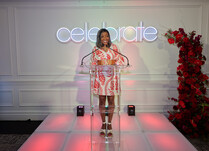 A woman in front of a "celebrate" neon sign and behind a podium addressing attendees